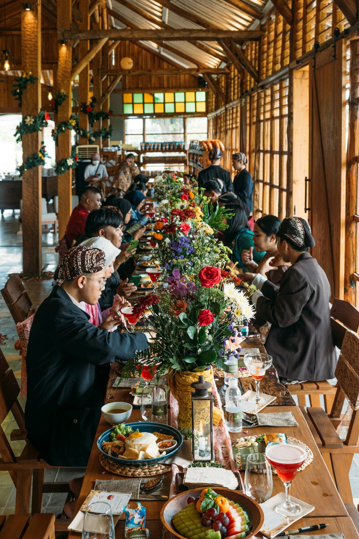A long table of guests at dinner in a timbered hall, flowers down the middle